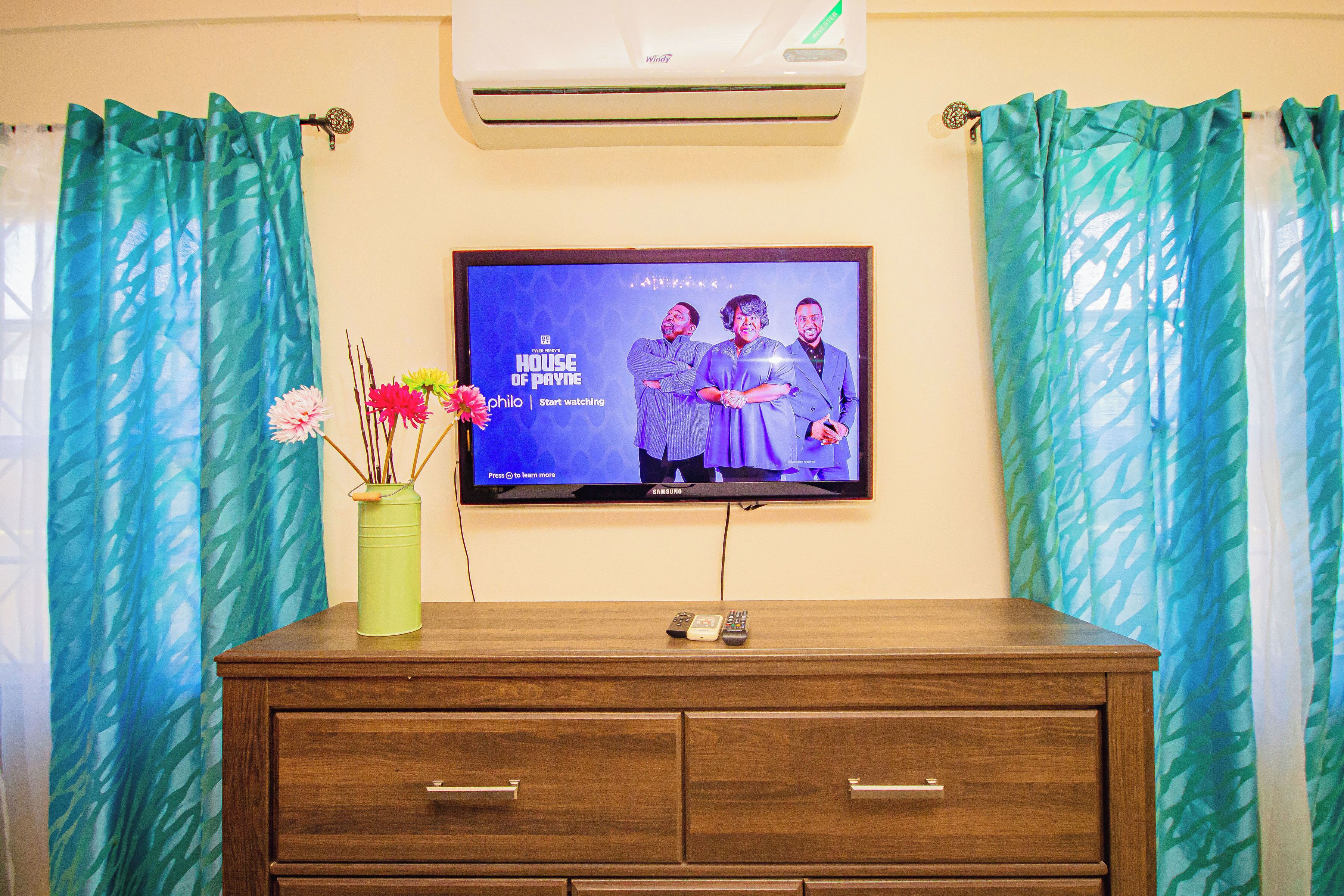 A television mounted on a wall displaying 'House of Payne' with a wooden dresser below.