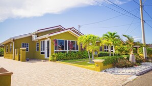 A yellow house with a paved driveway and a small garden with palm trees and shrubs. - Adorable Beach House In Draxhall (Ocho Rios)