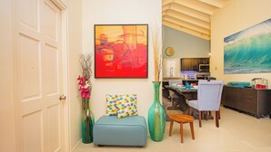 A living room with a sofa, a dining table, chairs, a large abstract painting, a vase with flowers, and a kitchen in the background. - Adorable Beach House In Draxhall (Ocho Rios)