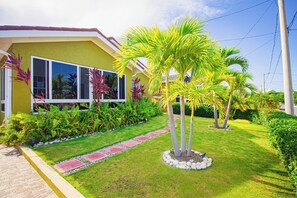 A yellow house with a well-maintained garden, including palm trees and a paved pathway. - Adorable Beach House In Draxhall (Ocho Rios)