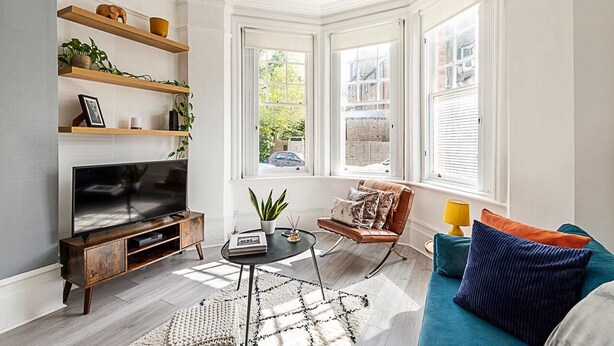 A modern living room with a large TV, wooden shelves, a sofa, and a window with a view of the street. - Stylish Hideaway Flat at Milton Park Highgate (London)