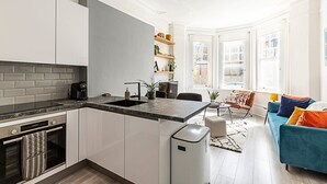 A modern kitchen with white cabinets, a black countertop, and a built-in oven. There is a dining area with a blue sofa and a small table. - Stylish Hideaway Flat at Milton Park Highgate (London)