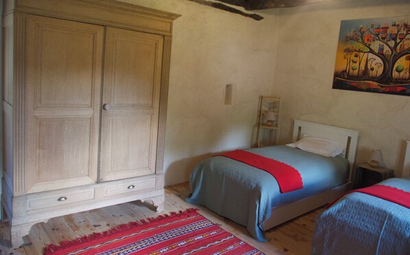 A bedroom with a large wooden wardrobe, two single beds with red and blue bedding, a wooden floor, and a painting on the wall.