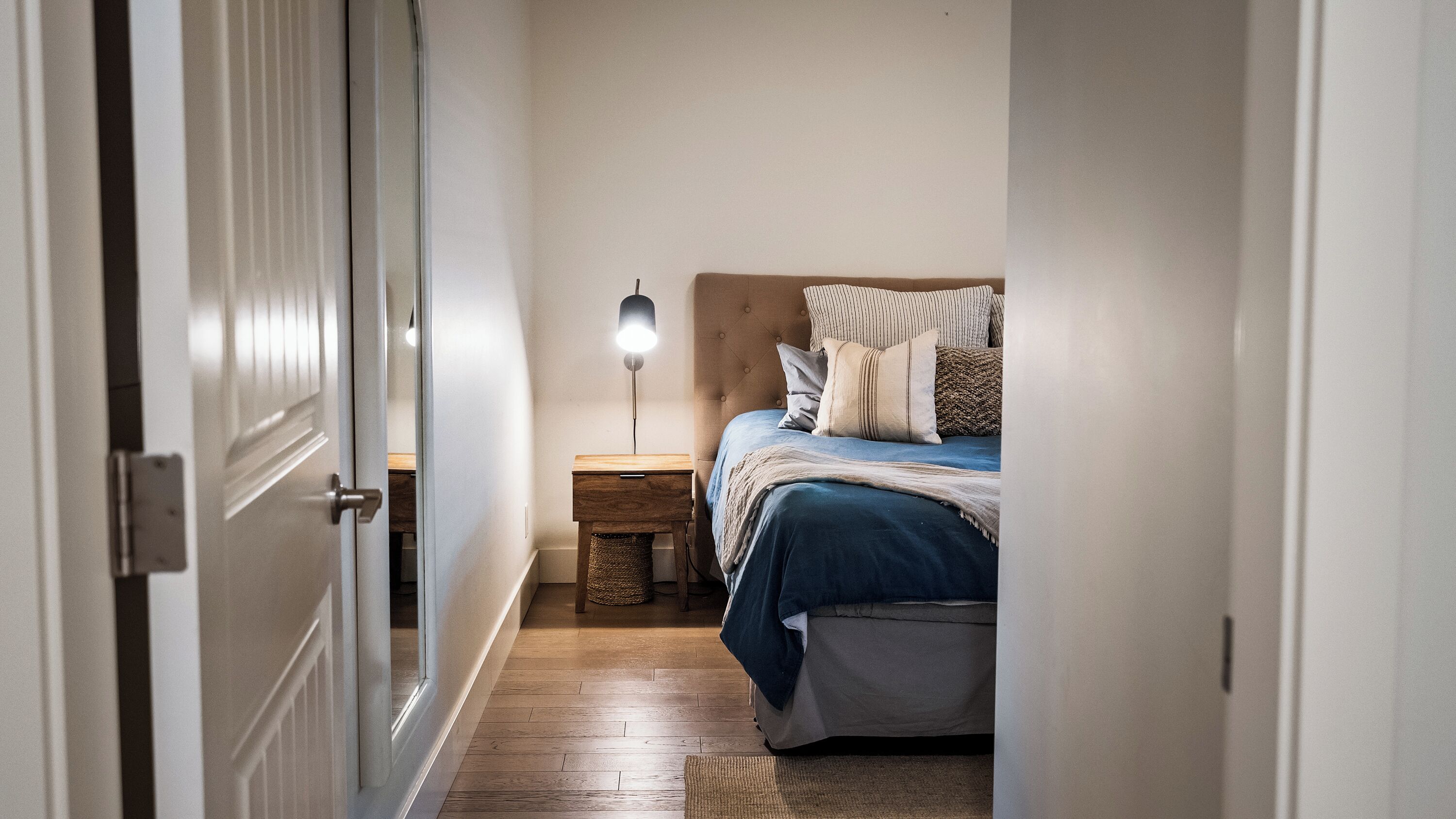 Un couloir étroit mène à une chambre à coucher qui contient un lit, une table de chevet, une lampe et une petite poubelle.