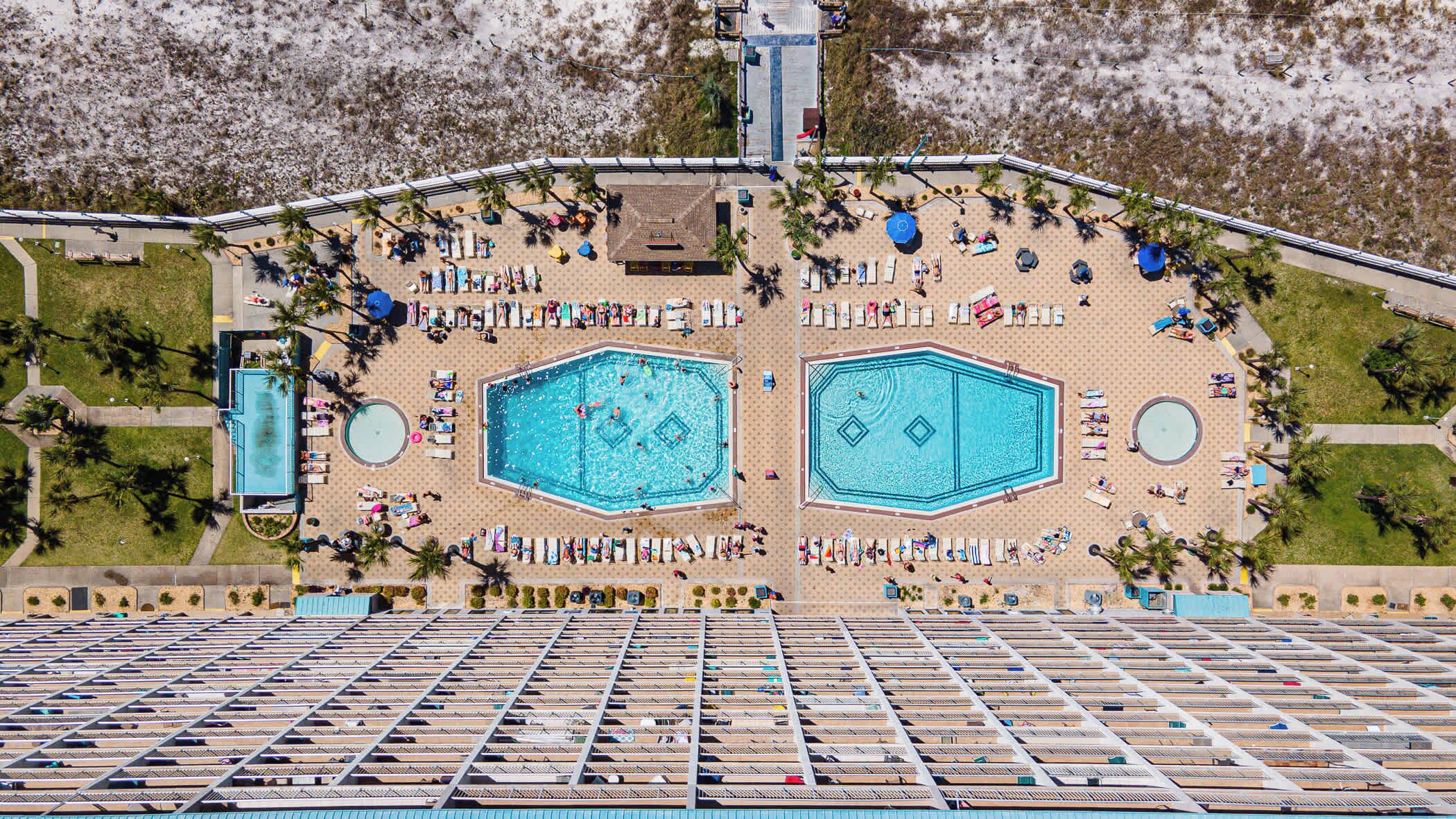An aerial view of a resort with two large swimming pools, a covered parking area, and a beach with sun loungers.