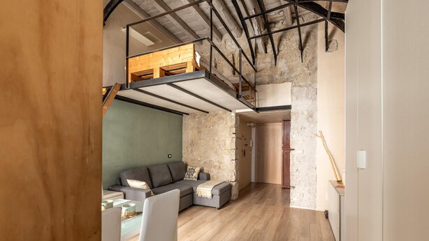 Un espace de vie de style loft avec une mezzanine en bois, un canapé gris et un mur en pierre.