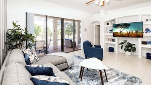 A spacious living room with a large sofa, a coffee table, and a TV mounted on the wall. There is a ceiling fan, a large window, and a door leading to an outdoor area. - Home Away From Home At Bridgeford Crossing (Davenport)