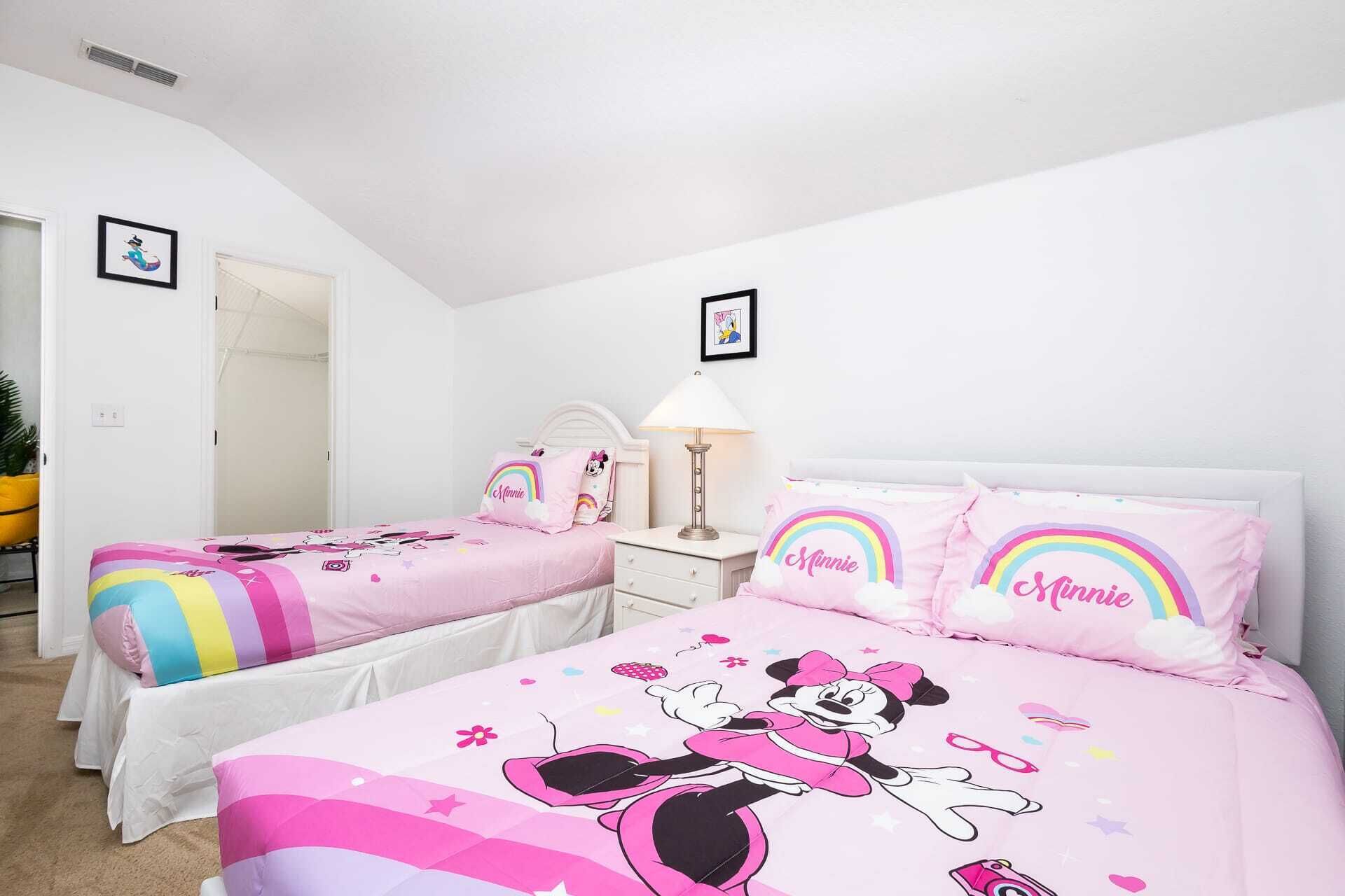 Et soveværelse med to senge, begge med pink sengetøj med Mickey Mus og regnbue-motiver. Der er et hvidt natbord med en lampe imellem sengene.