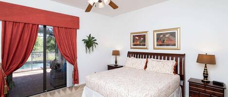 A bedroom with a bed, two nightstands, a ceiling fan, a large window with curtains, and two framed pictures on the wall.