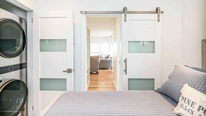 A bedroom with a bed, a sliding door leading to a living area, and a wall-mounted washing machine. - Latitude Adjustment Walk to Beach & Dining! (Scarborough)