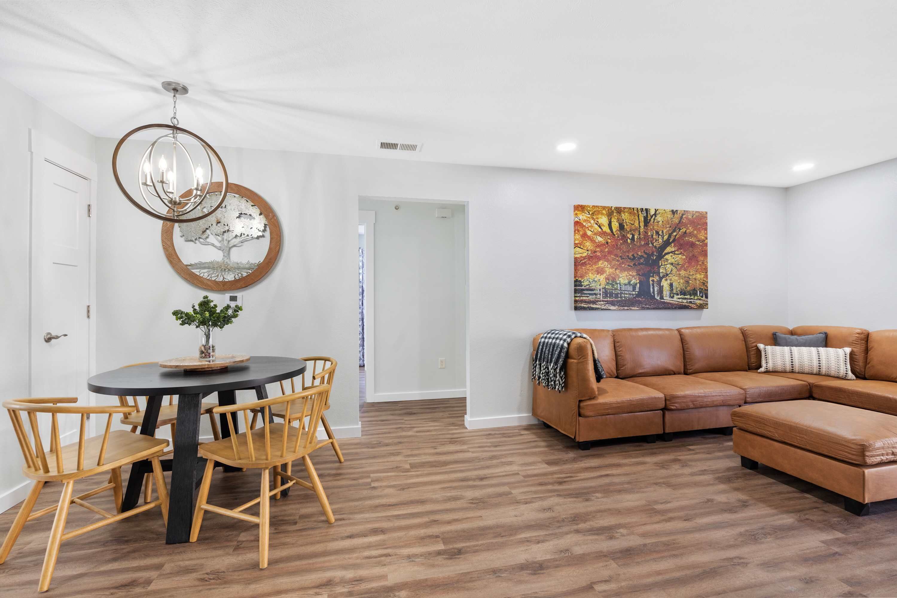 Un salon moderne avec une table à manger ronde, des chaises en bois, un canapé en cuir marron et un grand tableau sur le mur dont le thème est l’automne.