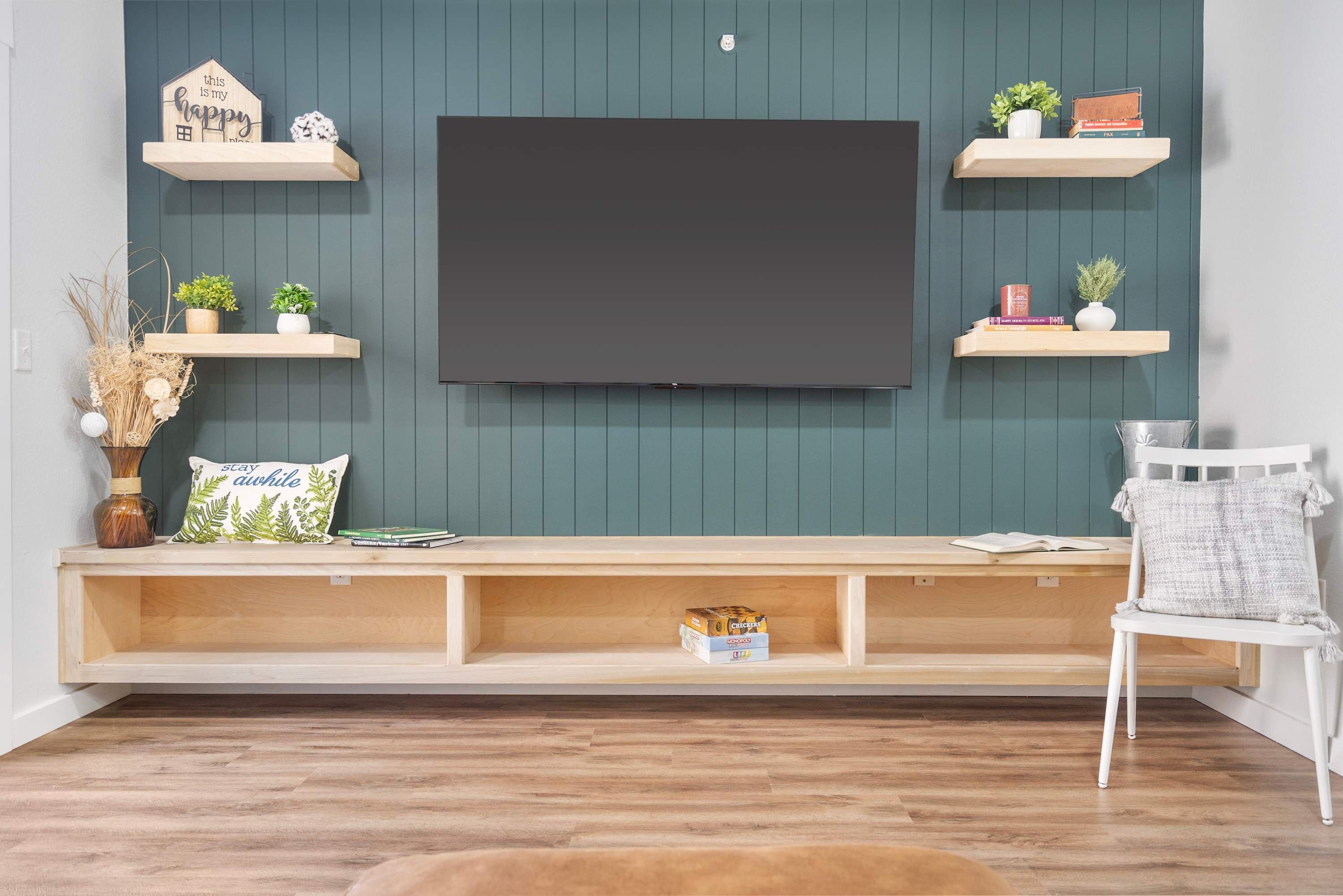 Un salon avec un téléviseur fixé au mur, des étagères en bois ornées de livres et de plantes, un banc avec espace de rangement et une chaise blanche.