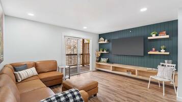 Un salón con un sofá de cuero marrón, una silla blanca, una mesa de centro de madera y un televisor montado en una pared verde oscuro.