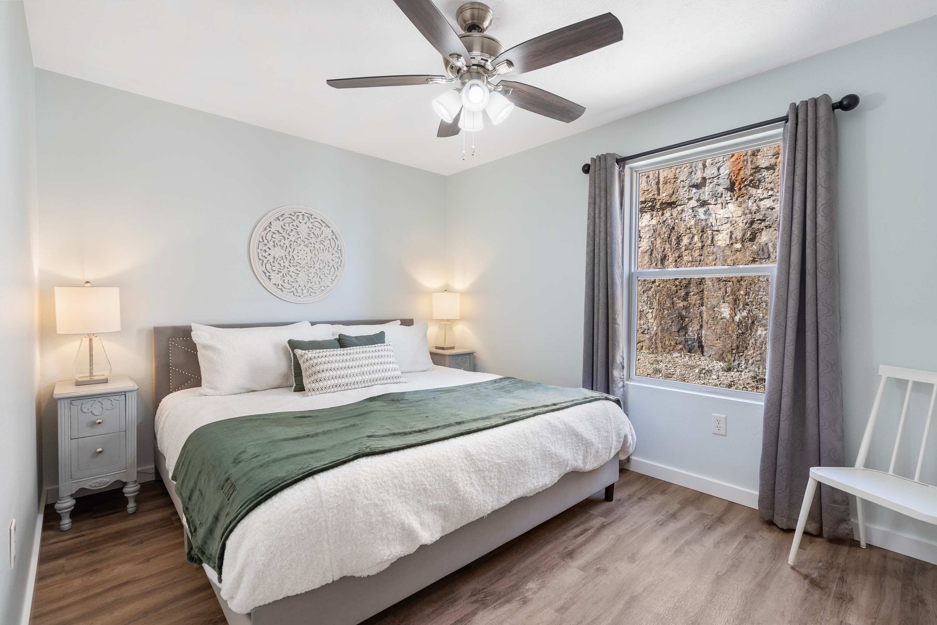 A bedroom with a bed, bedside lamps, a ceiling fan, a window with curtains, and a wall decoration.