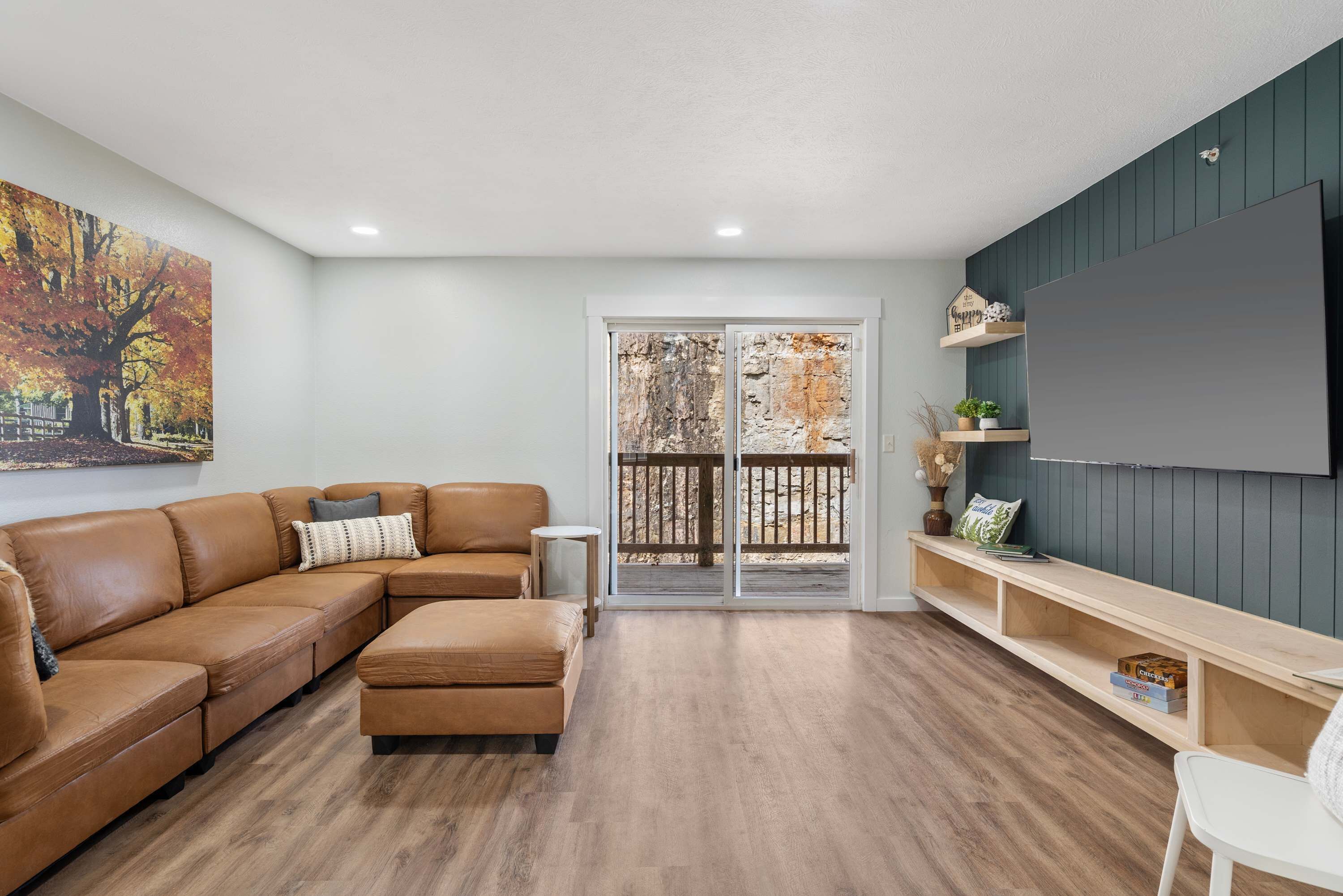 A living room with a brown sectional sofa, a small white table, and a wooden bench. There is a large flat-screen TV mounted on a dark wall, and a sliding glass door leading to an outdoor area.