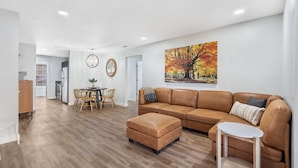 Ein modernes Wohnzimmer mit einer braunen Ledersofa, dazu passendem Hocker und einem Essbereich mit Holzsesseln und einem Tisch.