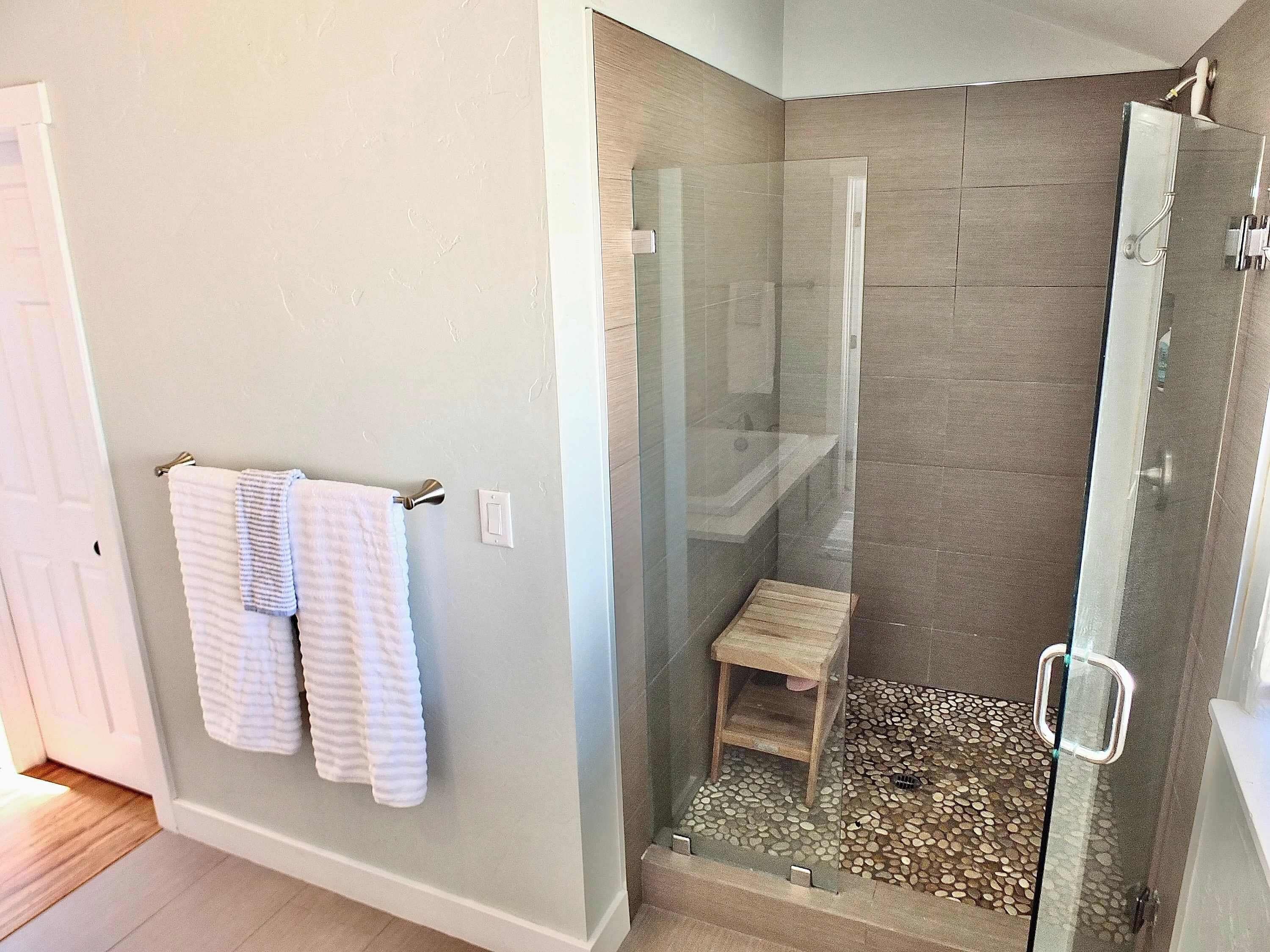 Een moderne badkamer met een douchecabine van glas, een houten kruk en een handdoekrek met twee handdoeken.