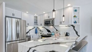 Una cucina moderna con mobili bianchi, elettrodomestici in acciaio inossidabile, un'isola in marmo e illuminazione a sospensione.
