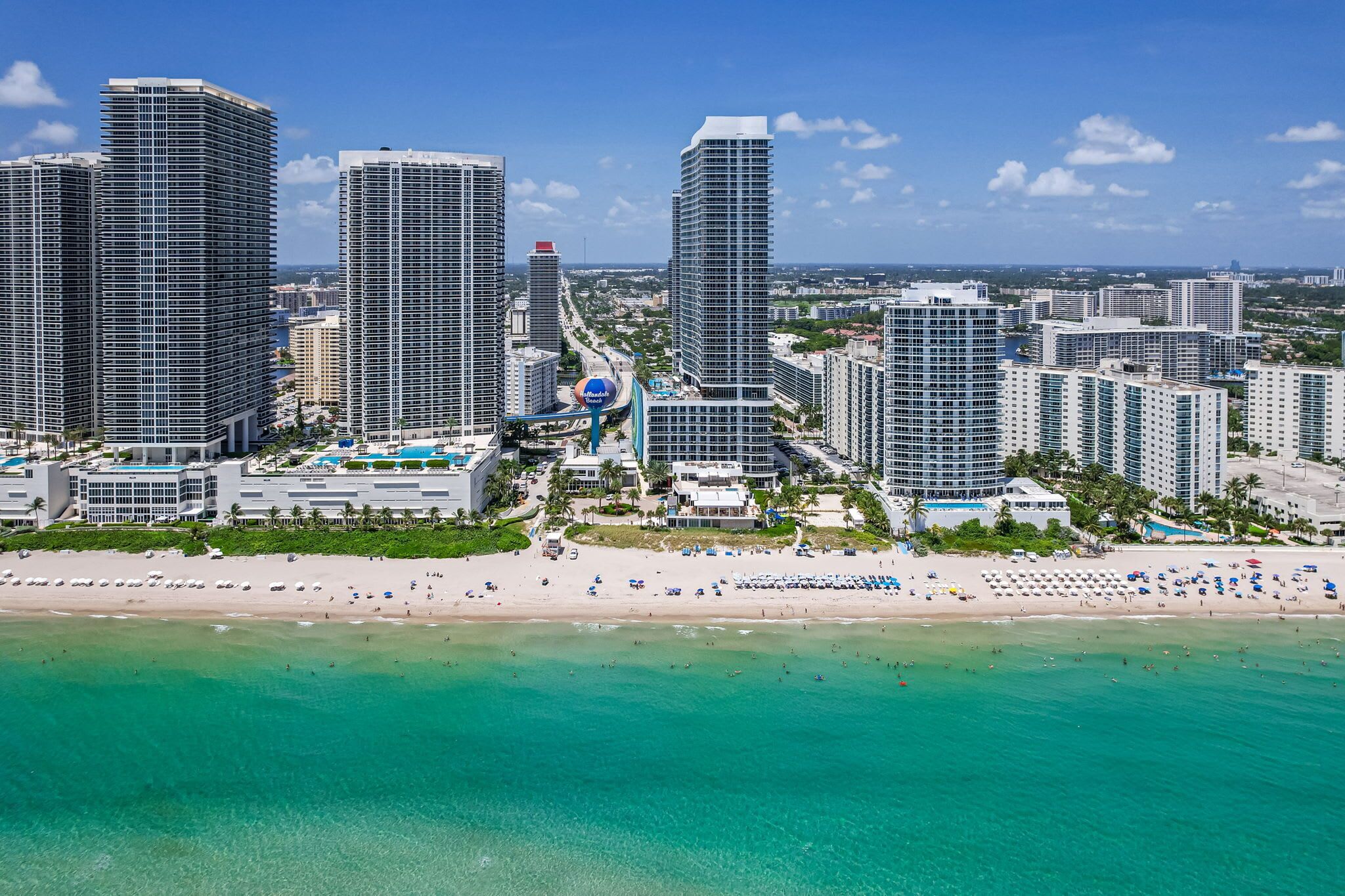 A coastal cityscape with high-rise buildings, a beach, and a clear blue sky.