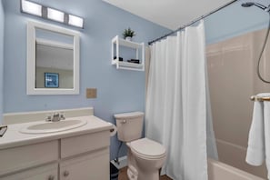 A bathroom with a white sink, toilet, and shower curtain. There is a mirror above the sink and a small shelf with a plant.
