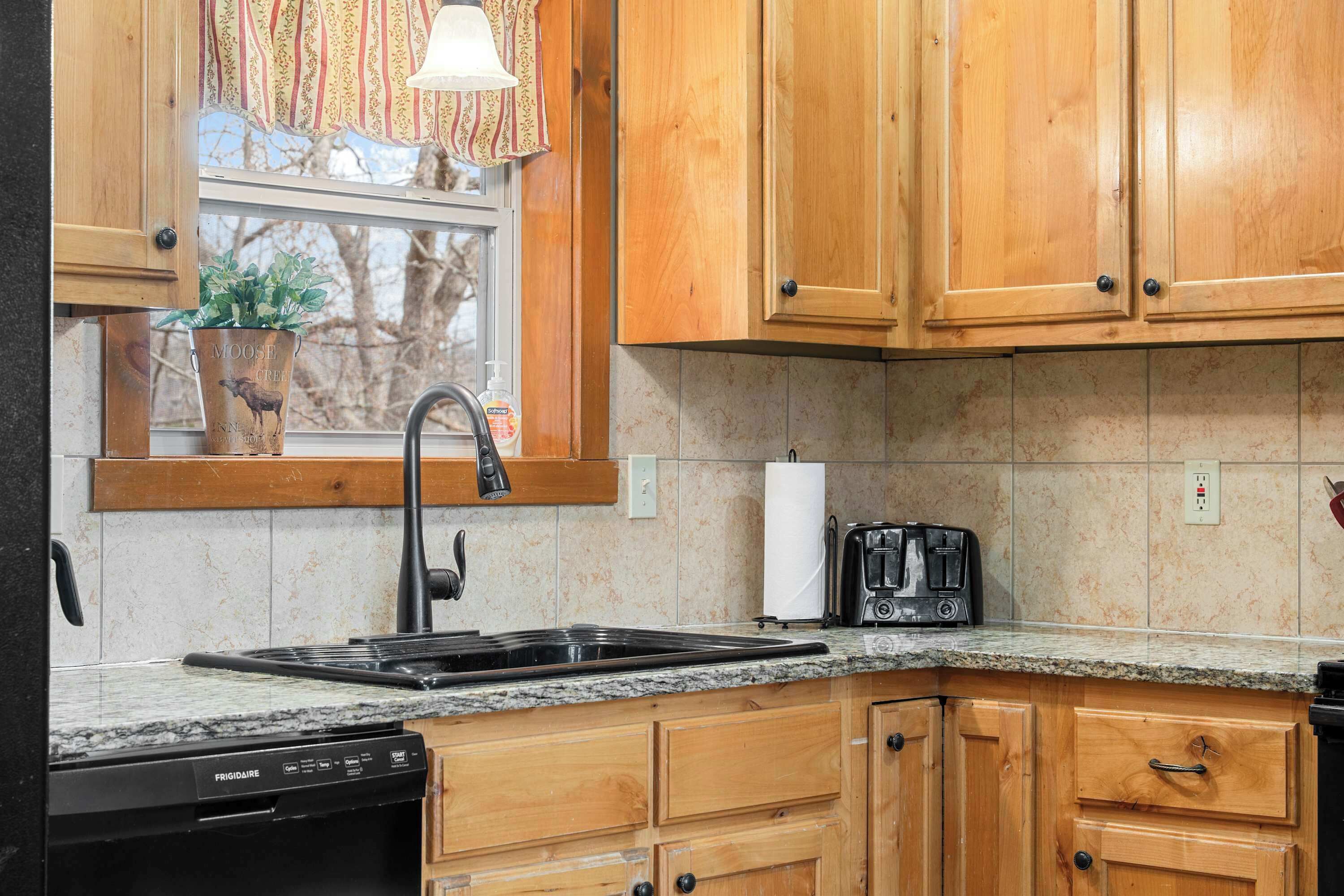 A kitchen with wooden cabinets, a granite countertop, a built-in dishwasher, and a window with a view of trees.