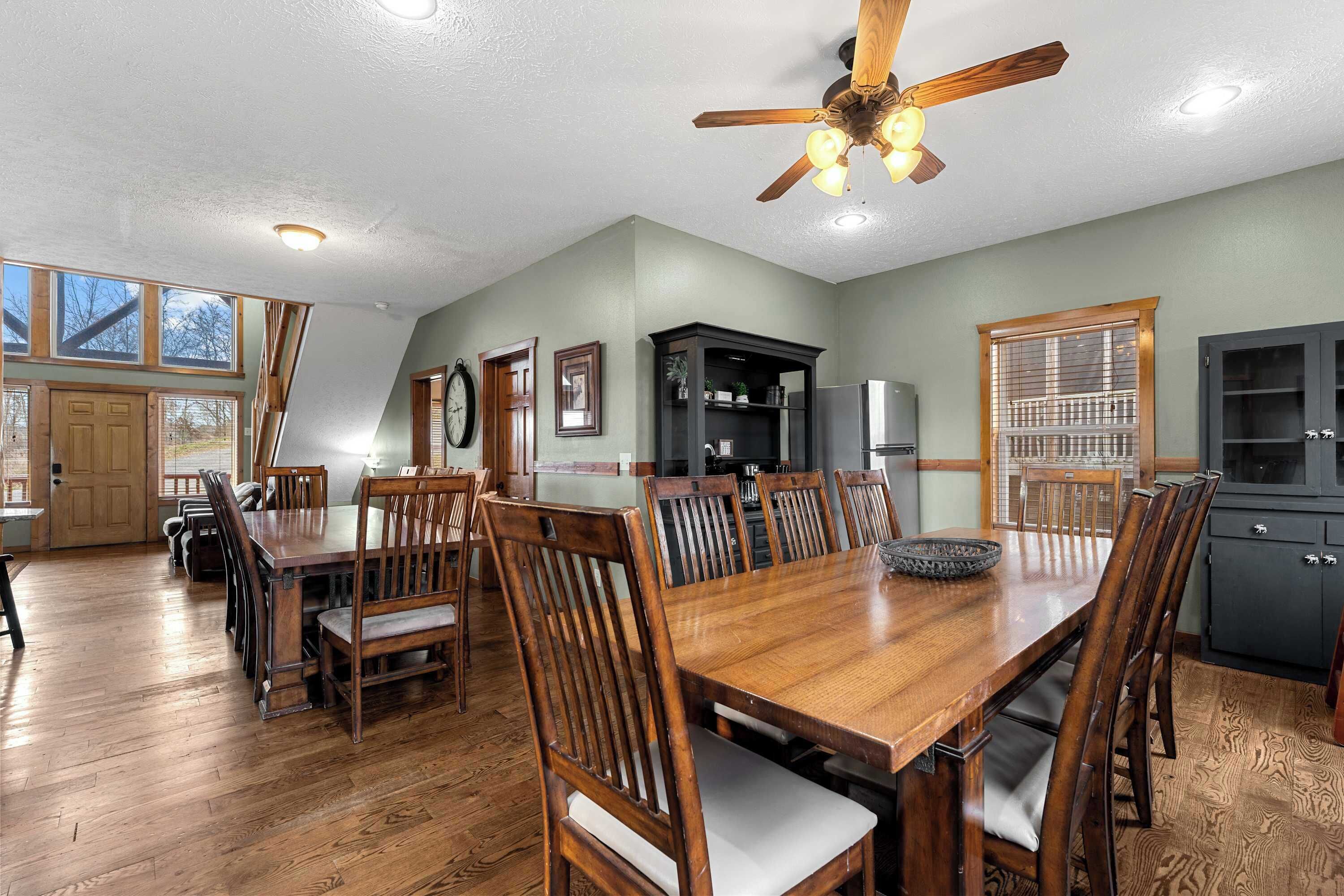 A spacious dining area with a wooden table and chairs, a ceiling fan, and a kitchen area with cabinets and a refrigerator.