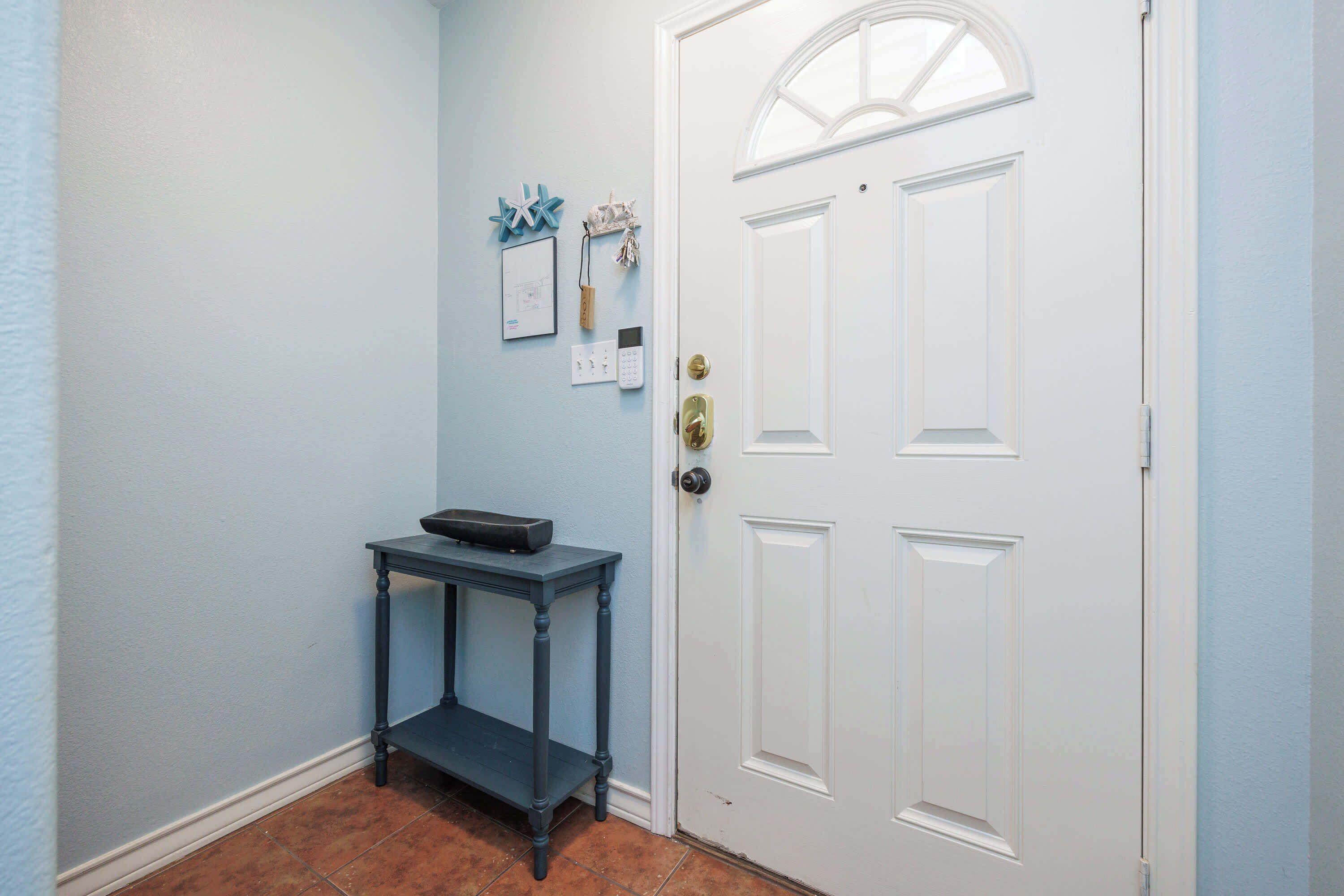 A hallway with a white door, a small table with a black object on it, and wall-mounted items including a light switch and a framed picture.