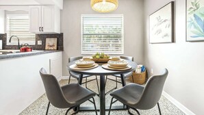 A modern kitchen with white cabinets, a round dining table with four chairs, a hanging light fixture, and a window with blinds. - Sunny Side Up A Charming Retreat! (Tampa)