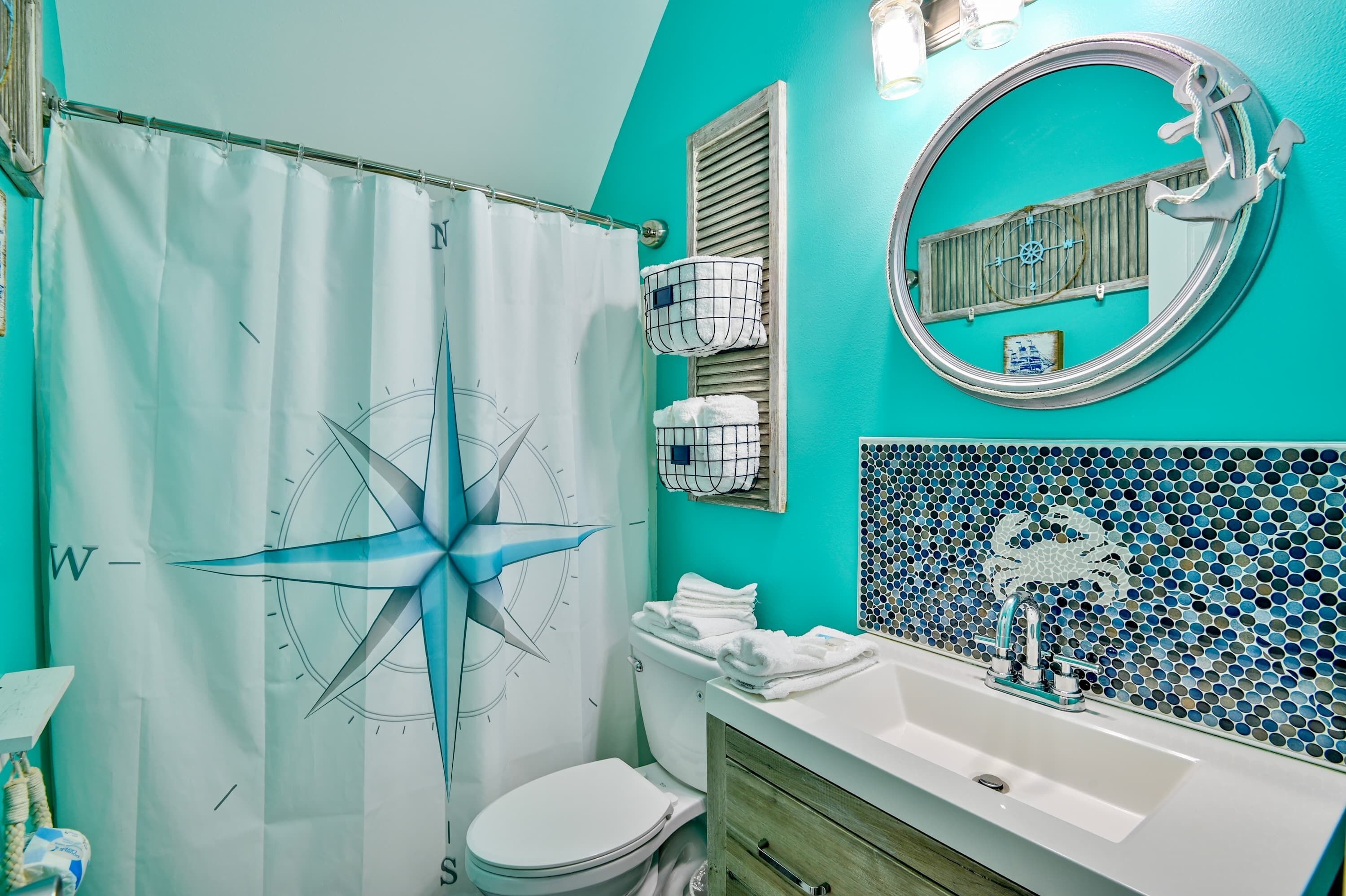 A bathroom with a nautical-themed shower curtain, a white toilet, a sink with a mirror, and a mosaic tile backsplash.