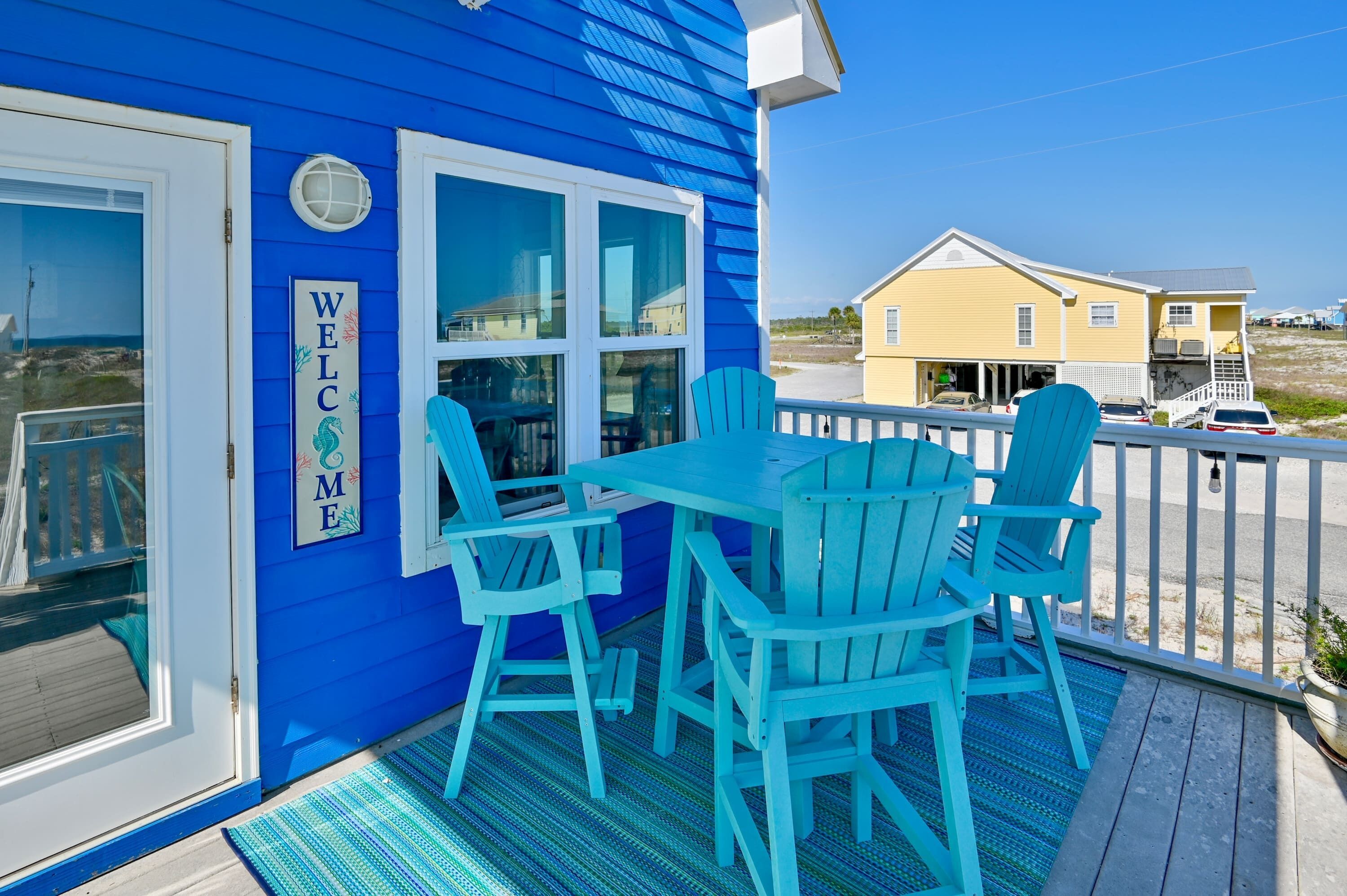 A blue building with a 'WELCOME' sign, a covered outdoor seating area with blue chairs, and a view of the beach and other buildings.