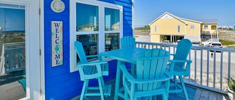 A blue building with a 'WELCOME' sign, a covered outdoor seating area with blue chairs, and a view of the beach and other buildings.