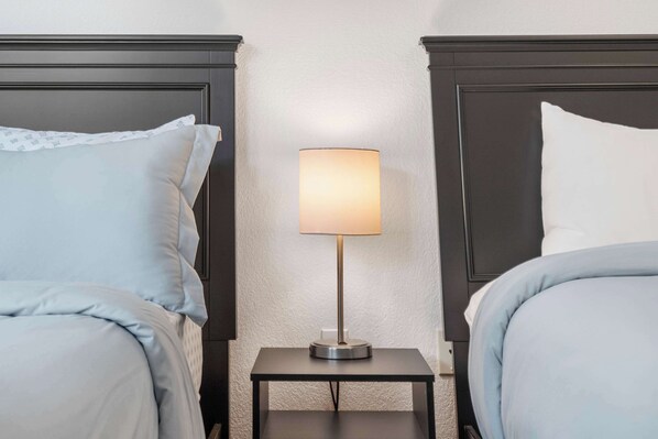 A hotel room with two beds, each with a pillow and a blanket. There is a small table with a lamp between the beds.