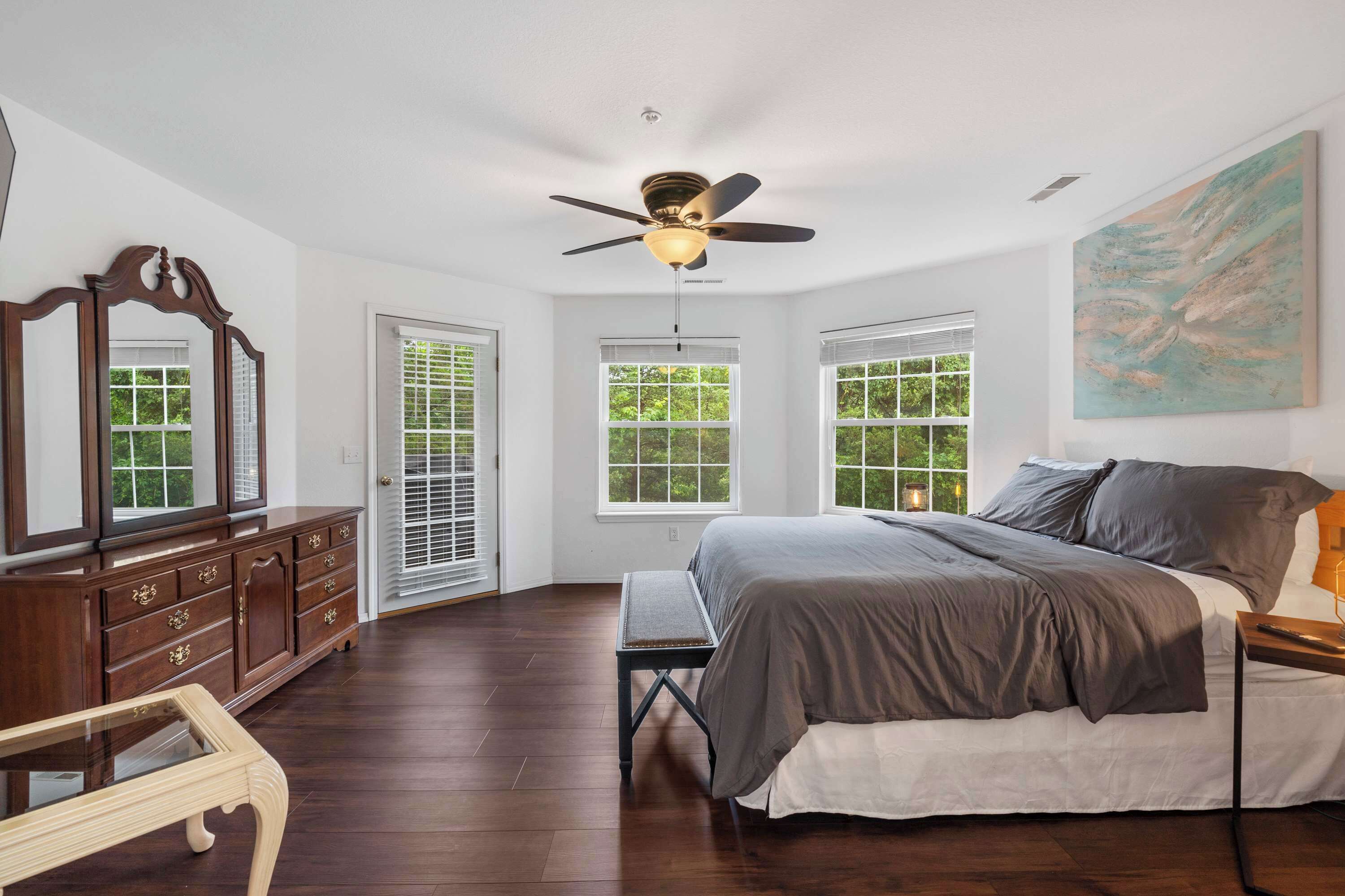 A bedroom with a large bed, a ceiling fan, a wooden dresser, and a window with blinds.