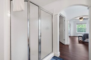 Un pasillo con puerta de vidrio para ducha, ventilador de techo y vista hacia un dormitorio con cama y ventana.