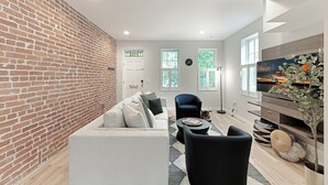 A modern living room with a brick wall, a sofa, a chair, a TV, and a plant. - Upscale Old Town Alexandria Retreat - Walk to King St Metro (Alexandria)