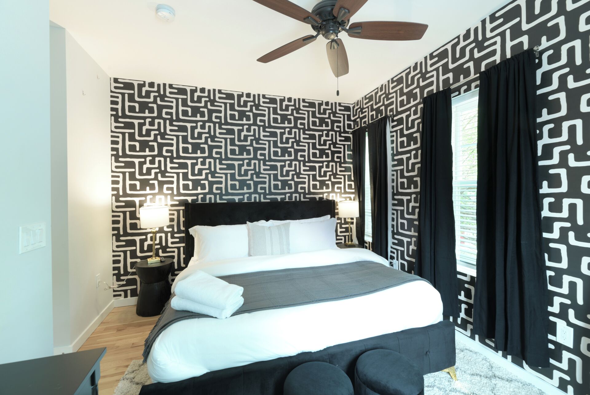Una moderna camera d'albergo con parete a motivo geometrico, una ventola a soffitto, un letto con biancheria bianca, un comodino con lampada e una finestra con tende nere.