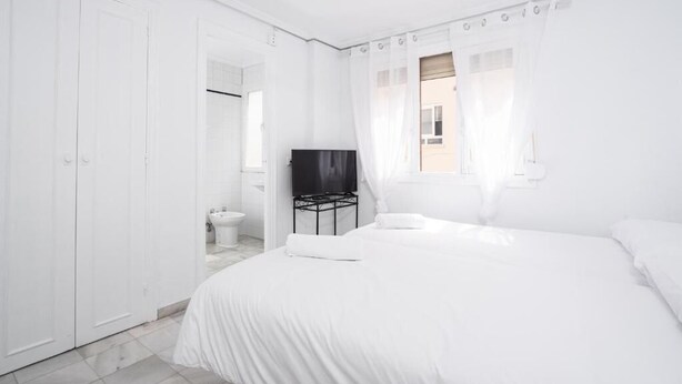 A white bedroom with a bed, a ceiling fan, a TV on a stand, and a window with curtains. - Hostal Alicia (Marbella)