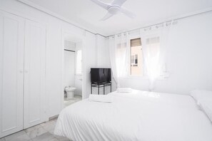 A white bedroom with a bed, a ceiling fan, a TV on a stand, and a window with curtains. - Hostal Alicia (Marbella)