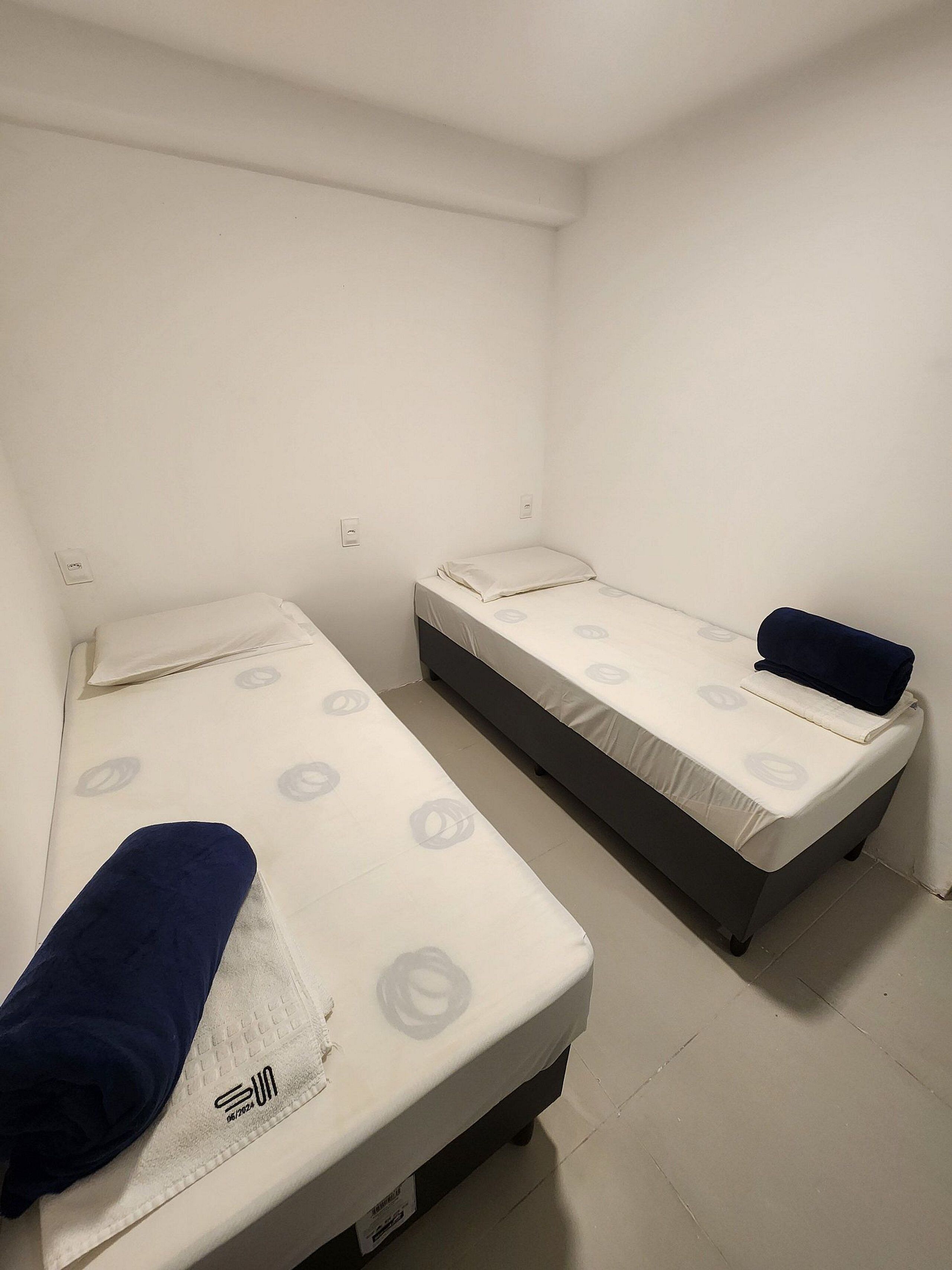 A small, clean room with two single beds, a white wall, and a visible electrical outlet.