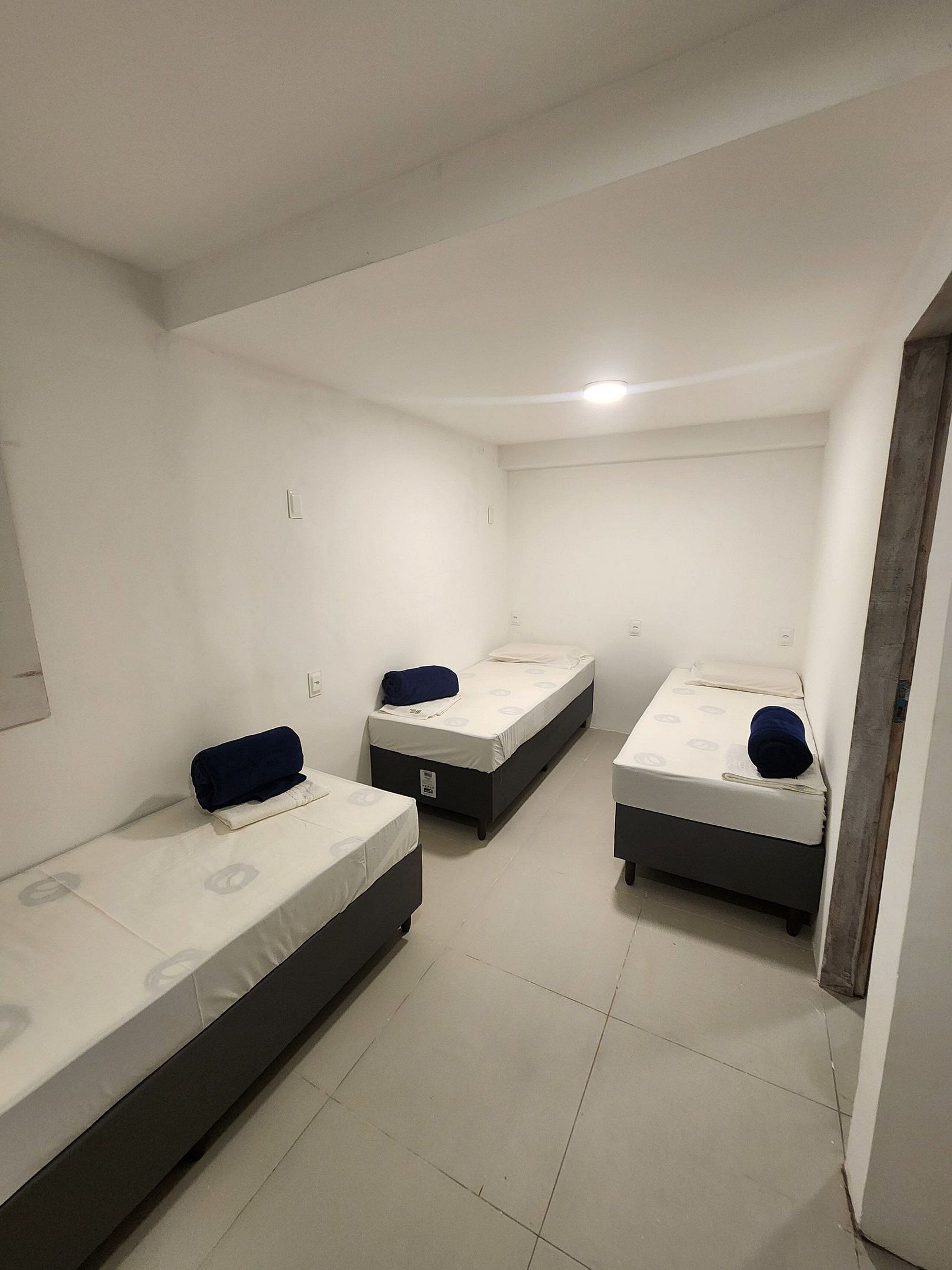 Una habitación con tres camas individuales, cada una con una almohada y una manta, paredes blancas y suelo de baldosas.