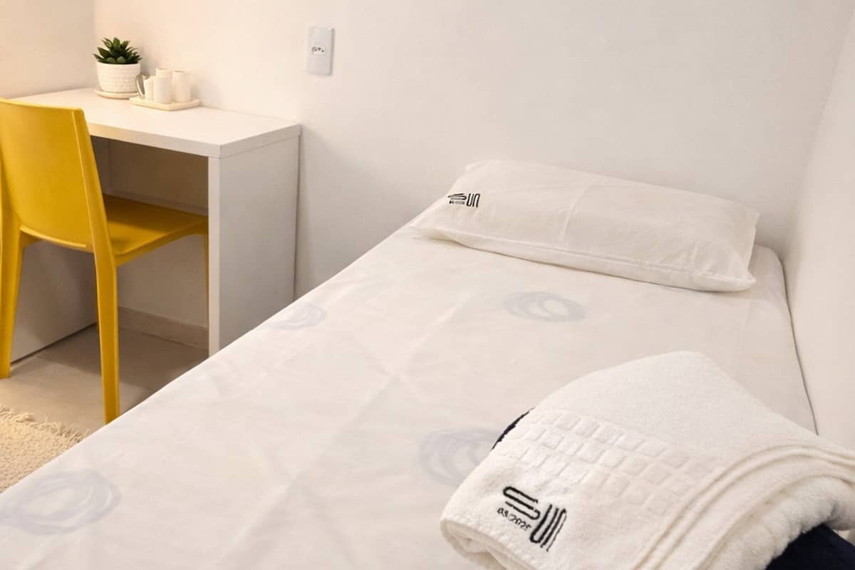 A single bed with a white blanket, a pillow, and a folded towel. There is a desk with a chair and a small potted plant. A framed picture hangs on the wall.
