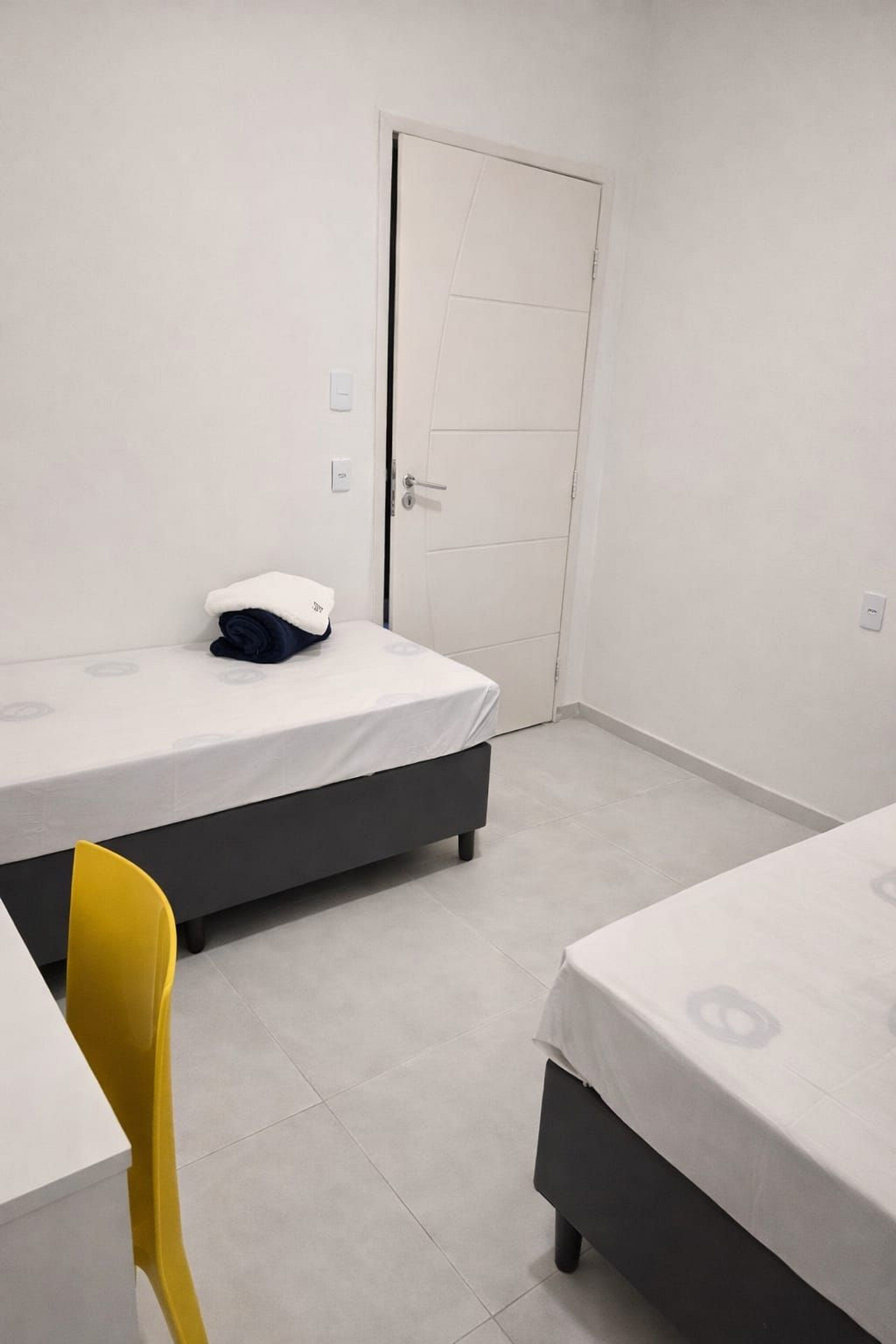 A small, clean room with two beds, a white door, and a yellow chair.