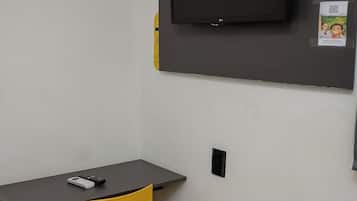 Quarto com cama, escrivaninha, cadeira, televisão fixada na parede e um pequeno cartaz.