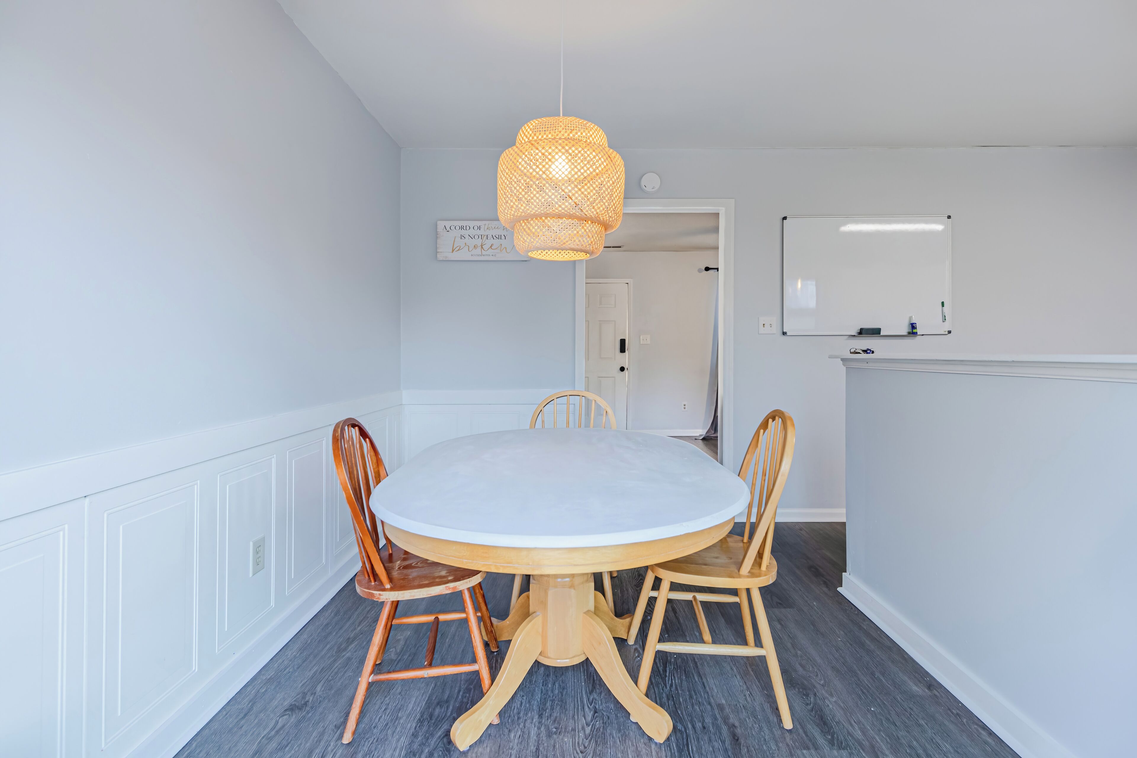 Un coin repas avec une table ronde et quatre chaises en bois, un luminaire suspendu en fibres tressées, un tableau blanc et une porte donnant sur une autre pièce.