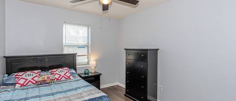 Ein Schlafzimmer mit Bett, Nachttisch, Kommode, Deckenventilator und Fenster mit Jalousien.