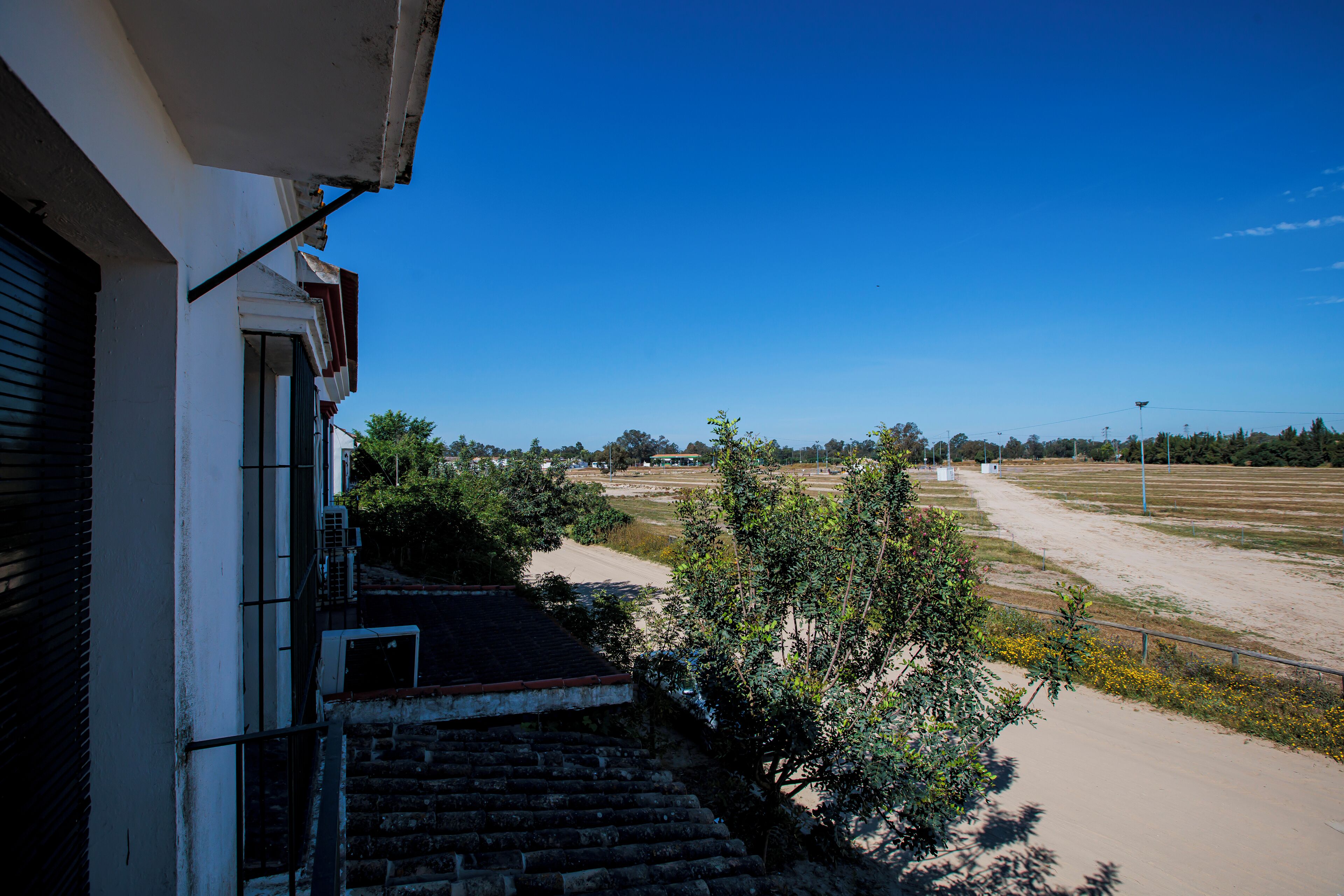 A residential building with a visible staircase leading to a balcony, a clear blue sky, and a dirt path in the background.