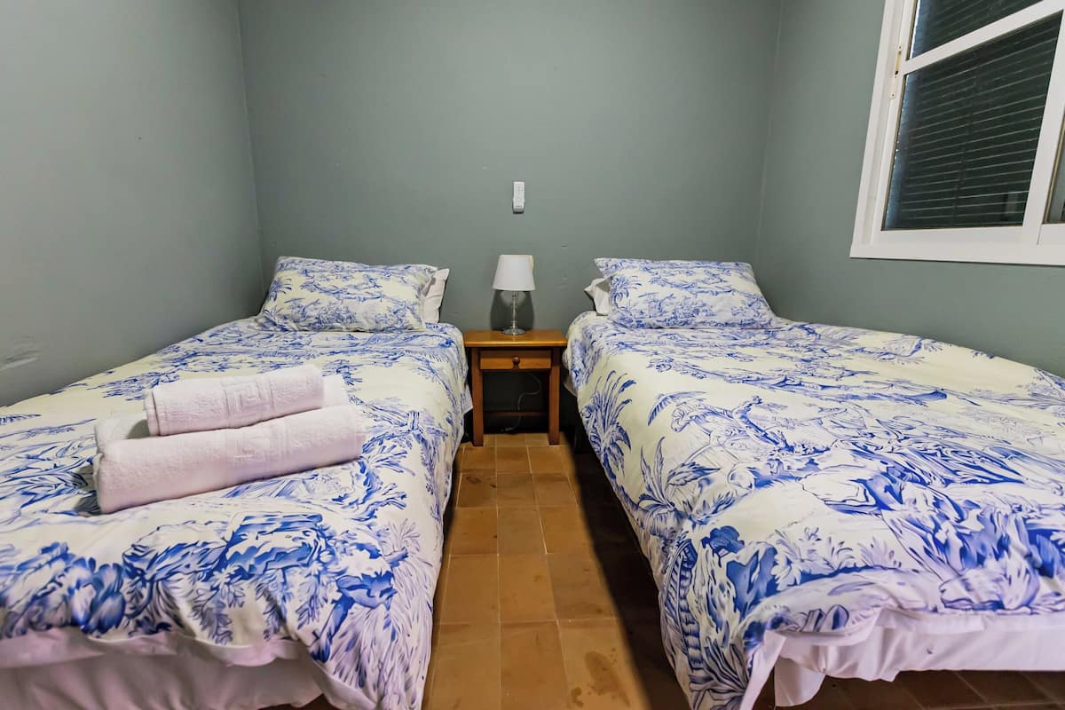 Habitación con dos camas, cada una con ropa de cama estampada con motivos florales, una mesita de noche pequeña con lámpara y una ventana con persianas.