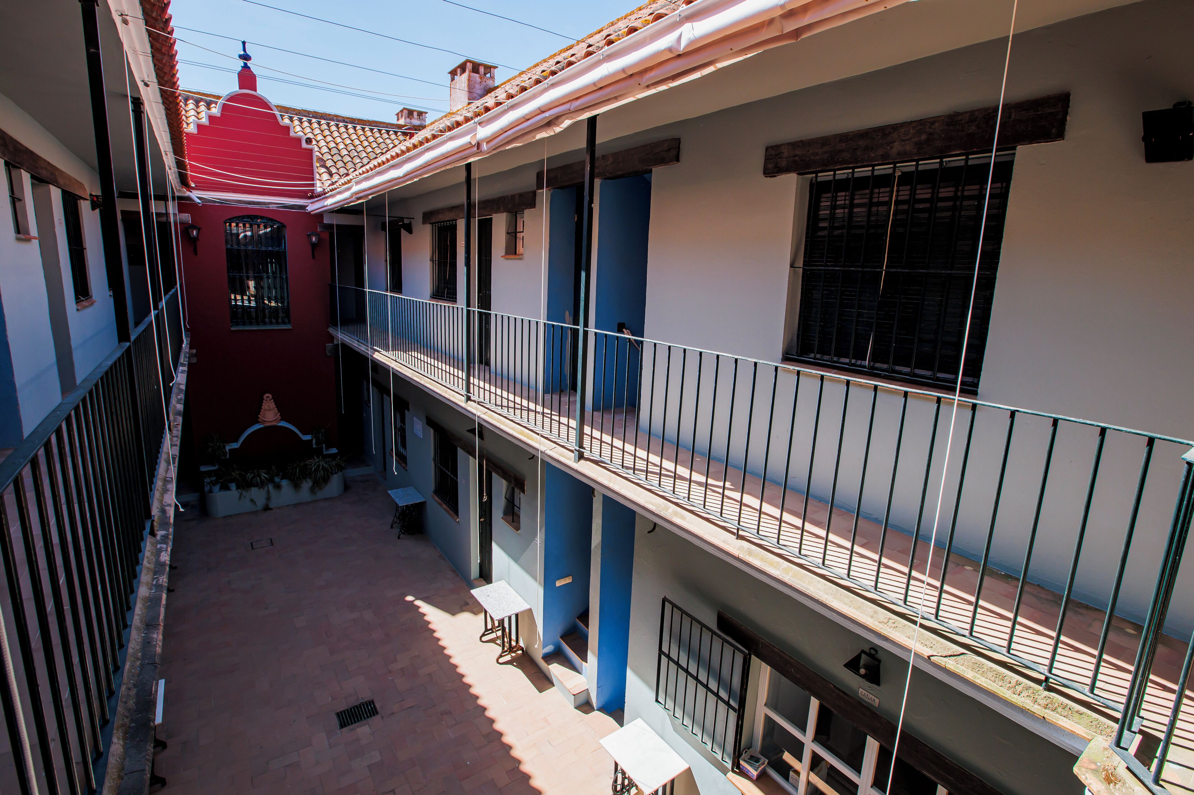 Un patio con un edificio rojo, pilares azules y un balcón con barandilla de metal.