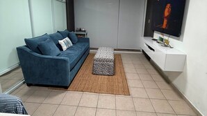 Un salon moderne avec un canapé bleu, un pouf à motifs, un téléviseur à écran plat et un meuble de rangement blanc pour les appareils multimédias.
