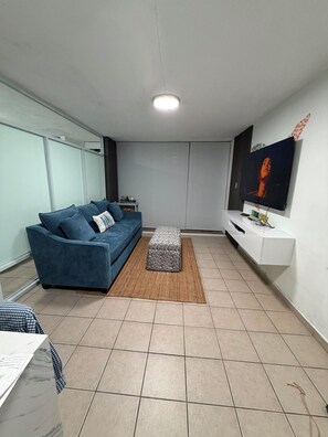 A modern living room with a blue sofa, a patterned ottoman, a flat-screen TV, and a white entertainment unit. - One minute walk to the beach! (Luquillo)